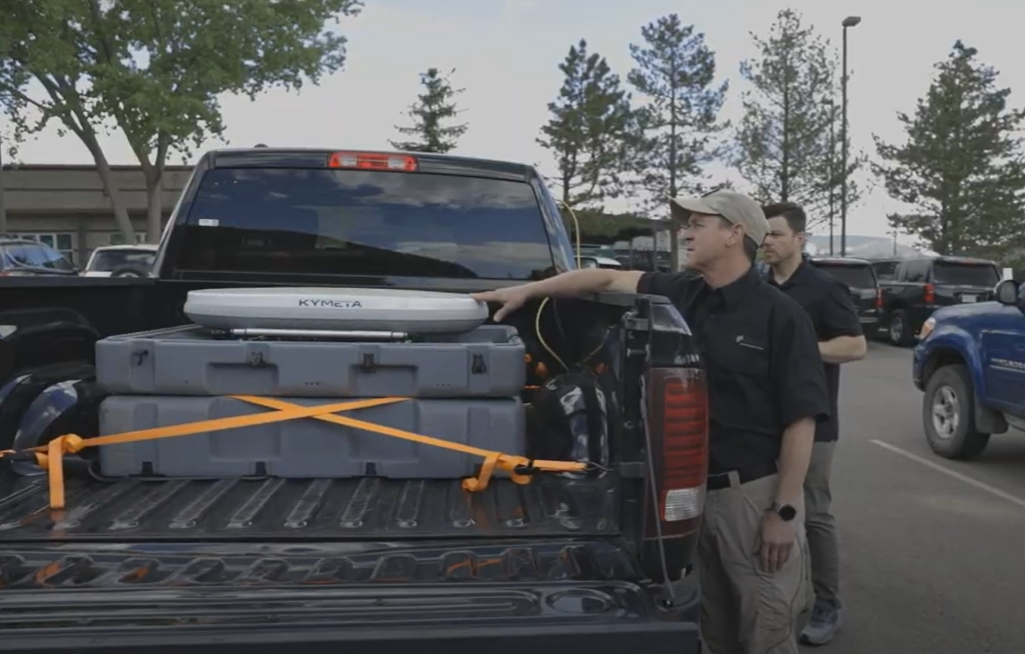 Truck bed carrying a Kymeta flat panel satellite antenna and secured equipment cases, with two individuals inspecting the setup for mobile satellite communication deployment.